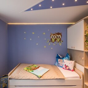 Modernes Kinderzimmer mit gemütlichem Bett, Wandtattoo Eulen, Pflanzen und Kuscheltieren.
