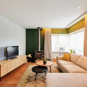 Modernes Wohnzimmer mit beiger Eckcouch, Holzofen, TV und grüner Akzentwand.